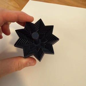 May include: A dark blue, star-shaped cookie cutter with multiple layers. The cutter has a central cylindrical handle. The object is held against a white surface, with a wooden surface visible in the background.