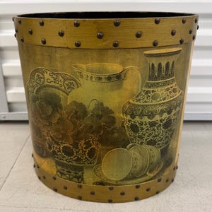 May include: A cylindrical wastebasket featuring a gold-toned rim and base accented with dark metal studs. The exterior surface displays a printed still life illustration of a pitcher, a tall vase, and teacups arranged with floral elements.