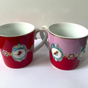 Puede incluir: Dos tazas de cerámica con interior blanco y bordes dorados. Una taza es de color rojo sólido, la otra es rosa y roja. Ambas presentan un medallón ovalado azul claro con una ilustración de un pájaro, rodeado de motivos florales y detalles dorados.