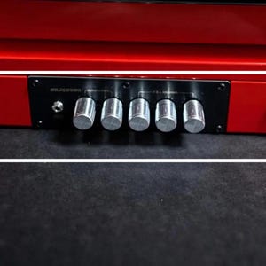 May include: A red electronic device with a black control panel featuring five silver knobs, a small input jack, and a red power button. The device is set against a dark background.