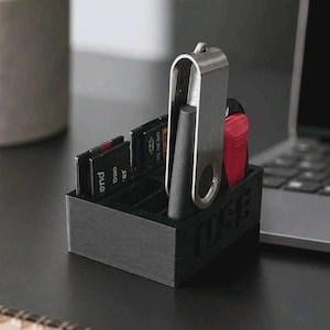 Può includere: Un organizer nero stampato in 3D con diversi scomparti che contiene varie schede di memoria SD e una chiavetta USB in metallo argentato. La parte anteriore del contenitore rettangolare presenta il testo in rilievo TUSE in carattere grassetto.