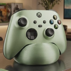 Può includere: Un controller wireless per videogiochi color verde salvia metallizzato appoggiato su una base di ricarica semicircolare abbinata. Il controller presenta levette analogiche nere, un pad direzionale nero e pulsanti azione colorati Y, B, A e X.
