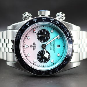 Puede incluir: Un reloj cron&oacute;grafo de acero inoxidable con una esfera degradada que va del rosa claro al azul claro. El reloj cuenta con un bisel taquim&eacute;trico negro, dos subesferas negras, una ventana de fecha y una pulsera de eslabones plateada. El texto en la esfera dice Tudor Geneve y Blaken 200m 660ft Chronometer Officially Certified.