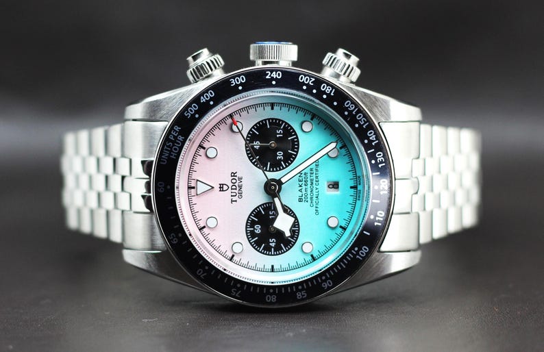 Puede incluir: Un reloj cron&oacute;grafo de acero inoxidable con una esfera degradada que va del rosa claro al azul claro. El reloj cuenta con un bisel taquim&eacute;trico negro, dos subesferas negras, una ventana de fecha y una pulsera de eslabones plateada. El texto en la esfera dice Tudor Geneve y Blaken 200m 660ft Chronometer Officially Certified.