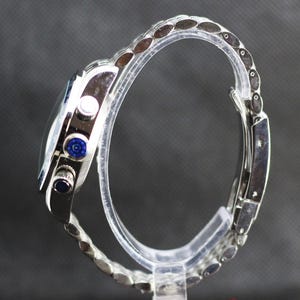 Puede incluir: Un reloj cron&oacute;grafo de tono plateado con brazalete de eslabones met&aacute;licos, caja circular y tres botones laterales. El bot&oacute;n central tiene un patr&oacute;n circular azul, mientras que el bot&oacute;n inferior presenta una incrustaci&oacute;n circular azul oscuro.
