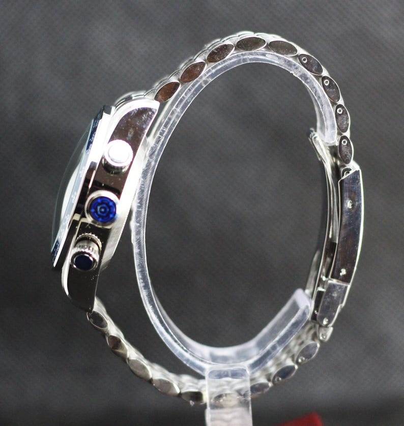 Puede incluir: Un reloj cron&oacute;grafo de tono plateado con brazalete de eslabones met&aacute;licos, caja circular y tres botones laterales. El bot&oacute;n central tiene un patr&oacute;n circular azul, mientras que el bot&oacute;n inferior presenta una incrustaci&oacute;n circular azul oscuro.