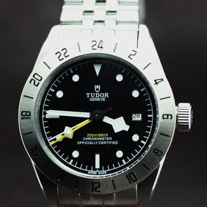 Könnte beinhalten: Eine Armbanduhr aus Edelstahl mit schwarzem Zifferblatt, weißen Zeigern und Datumsanzeige. Auf dem Zifferblatt steht "TUDOR GENEVE" und "200m:660ft CHRONOMETER OFFICIALLY CERTIFIED". Das Armband ist ebenfalls aus Edelstahl.