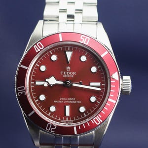 Pode incluir: Um relógio de pulso em aço inoxidável com uma moldura vermelha e um mostrador vermelho a condizer. O mostrador do relógio apresenta marcadores de horas e ponteiros brancos, com a marca "TUDOR GENEVE" visível. A pulseira é em aço inoxidável.