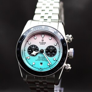 Puede incluir: Un reloj cron&oacute;grafo de acero inoxidable con brazalete de eslabones plateado. La esfera presenta un degradado de rosa a turquesa, subesferas negras, ventana de fecha y el texto Tudor Geneve y Blaken 200m:660ft Chronometer Officially Certified.