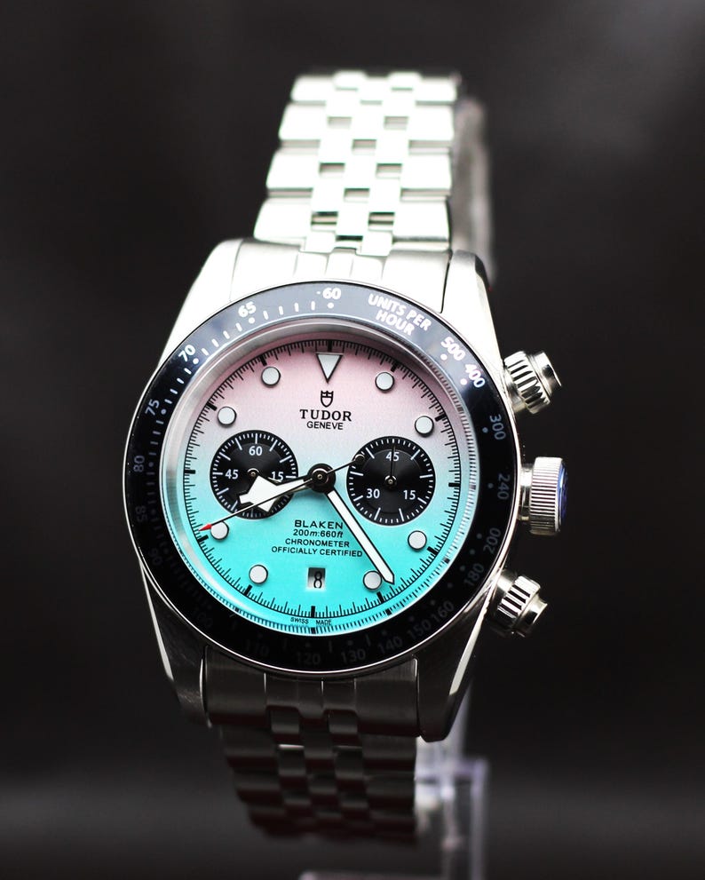 Puede incluir: Un reloj cron&oacute;grafo de acero inoxidable con brazalete de eslabones plateado. La esfera presenta un degradado de rosa a turquesa, subesferas negras, ventana de fecha y el texto Tudor Geneve y Blaken 200m:660ft Chronometer Officially Certified.