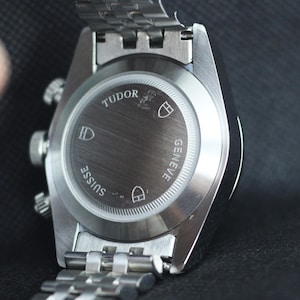 Puede incluir: Un reloj de pulsera de acero inoxidable en tono plateado con una correa de eslabones met&aacute;licos. La parte posterior de la caja es de metal cepillado y presenta el texto grabado TUDOR, GENEVE y SUISSE, junto con tres logotipos de escudos.