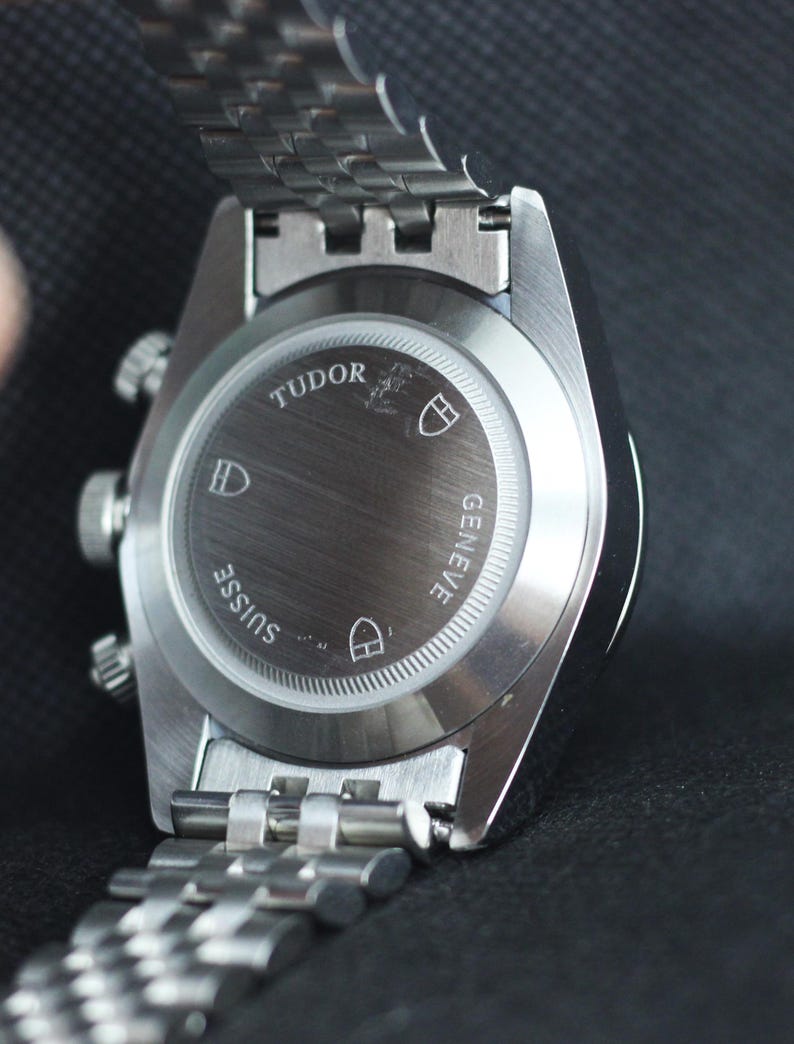 Puede incluir: Un reloj de pulsera de acero inoxidable en tono plateado con una correa de eslabones met&aacute;licos. La parte posterior de la caja es de metal cepillado y presenta el texto grabado TUDOR, GENEVE y SUISSE, junto con tres logotipos de escudos.