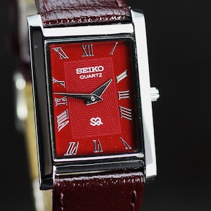 Peut inclure: Une montre Seiko Quartz rectangulaire avec un bracelet en cuir bordeaux. Le cadran de la montre est rouge avec des chiffres romains et des aiguilles argentés. Le boîtier est argenté et le bracelet en cuir est texturé.