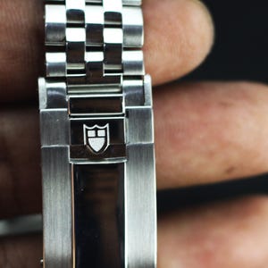 Puede incluir: Una correa de reloj de acero inoxidable con acabado de metal cepillado y un cierre central pulido. El cierre est&aacute; grabado con un logotipo de escudo blanco que contiene una l&iacute;nea horizontal y una vertical, el emblema de la marca Tudor.