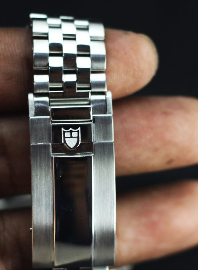 Puede incluir: Una correa de reloj de acero inoxidable con acabado de metal cepillado y un cierre central pulido. El cierre est&aacute; grabado con un logotipo de escudo blanco que contiene una l&iacute;nea horizontal y una vertical, el emblema de la marca Tudor.