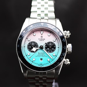 May include: A silver-tone stainless steel chronograph watch with a gradient dial transitioning from white at the top to turquoise at the bottom. It features a black tachymeter bezel, two black sub-dials, a date window, and a multi-link metal bracelet.