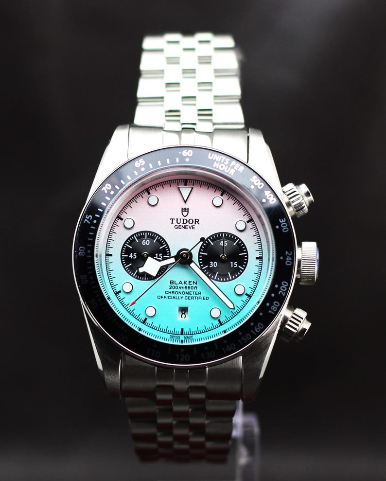 Puede incluir: Un reloj cron&oacute;grafo de acero inoxidable en tono plateado con una esfera degradada que va del blanco en la parte superior al turquesa en la inferior. Cuenta con un bisel taquim&eacute;trico negro, dos subesferas negras, una ventana de fecha y un brazalete de eslabones.