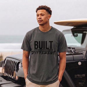 Peut inclure: T-shirt gris foncé avec les mots "BUILT DIFFERENT" en lettres noires. Le t-shirt est à manches courtes et a un col rond. Un Jeep noir et une planche de surf sont en arrière-plan.