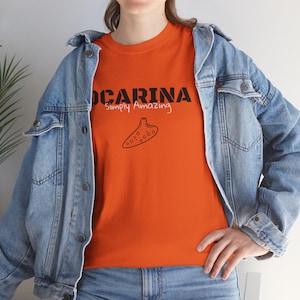 May include: An orange t-shirt with the word "OCARINA" in black block letters above the words "Simply Amazing" in a cursive font. A black line drawing of an ocarina is below the text. The person is wearing a denim jacket.