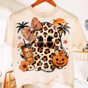 Puede incluir: Una camiseta color crema con el gráfico de un fantasma con estampado de leopardo, gafas de sol en forma de corazón y un lazo naranja brillante. El fantasma sostiene una bebida con la etiqueta Boozy & Beachy y un cubo de calabaza con frutas.