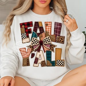 Può includere: Una felpa bianca a maniche lunghe con la scritta FALL Y'ALL in un design patchwork. Le lettere sono composte da vari motivi di tessuto, tra cui tartan, stampa leopardata e vichy, arricchite da piccoli bottoni e un fiocco in tessuto centrale.