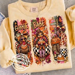 May include: A pale yellow crewneck sweatshirt featuring three vertical panels with Thanksgiving-themed graphics. The text reads GOBBLE BABE, CUTE, THANKFUL, FEAST, and ORIGINAL. The designs include turkeys, a pumpkin, leopard print boots, and a mug.