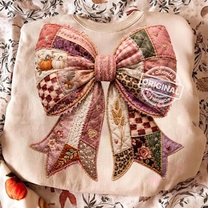 Pode incluir: Uma camisola de cor creme com um grande gráfico central de um laço em estilo acolchoado. O laço é composto por vários padrões de patchwork em tons de rosa, verde e castanho, incluindo abóboras, trigo, flores e corações. A palavra ORIGINAL está impressa.