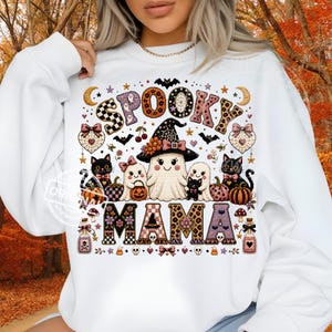 Puede incluir: Una sudadera blanca de manga larga con un estampado gráfico que incluye el texto SPOOKY MAMA. El diseño muestra un fantasma central con sombrero de bruja, rodeado de gatos negros, fantasmas pequeños, calabazas, murciélagos, estrellas y frascos.