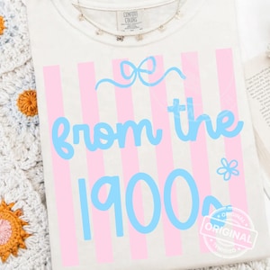 Puede incluir: Camiseta color crema con un diseño de rayas verticales rosas y las palabras "from the 1900s" en azul claro. Una pequeña flor y un lazo acentúan el texto. La camiseta está doblada y expuesta sobre una manta de ganchillo.