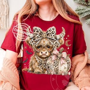 May include: A maroon t-shirt featuring a graphic of a Highland cow wearing heart-shaped leopard print glasses and a matching bow. The cow is adorned with a pearl necklace and sits beside a leopard print pumpkin with a pink bow, bats, and stars.