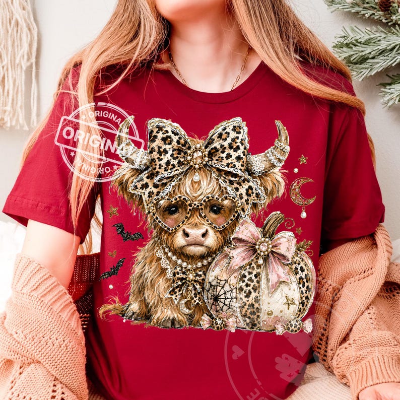 May include: A maroon t-shirt featuring a graphic of a Highland cow wearing heart-shaped leopard print glasses and a matching bow. The cow is adorned with a pearl necklace and sits beside a leopard print pumpkin with a pink bow, bats, and stars.