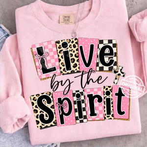Puede incluir: Sudadera rosa claro con la frase "Live by the Spirit" en una fuente decorativa. Las letras presentan varios patrones, incluyendo estampado de leopardo, lunares rosas y diseños a cuadros en blanco y negro. También se incluye un gráfico de cruz.