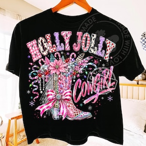May include: A black short-sleeve t-shirt featuring a graphic print of a pink cowboy boot decorated with a large bow, poinsettias, and ornaments. The text HOLLY JOLLY COWGIRL is printed in a stylized pink and white checkered font above and below.