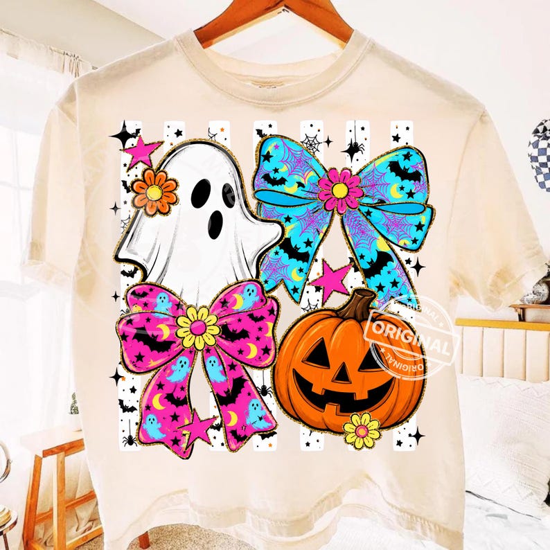 以下が含まれることがあります： ハロウィーンをテーマにしたデザインのクリーム色のTシャツ。デザインには、白い幽霊、コウモリとクモの巣が付いた2つのリボン、ジャック・オー・ランタン、星が含まれています。リボンはピンクと青で、黄色の花のアクセントが付いています。