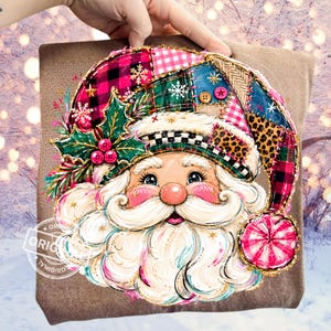 May include: A brown sweatshirt featuring a graphic of a Santa Claus face wearing a patchwork-style hat. The hat includes red and black buffalo plaid, pink gingham, and leopard print patterns, accented with green holly leaves and red berries.