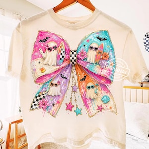 Pu&ograve; includere: T-shirt color crema con un grande disegno a fiocco colorato. Il fiocco presenta personaggi fantasma con occhiali da sole e tavole da surf. Il fiocco ha sezioni rosa, viola e turchese con stelle e conchiglie. La maglietta ha uno scollo tondo.