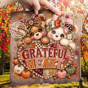 May include: A tan fabric item featuring two cartoon bears with large bows on their heads, positioned above a heart-shaped graphic containing the text GRATEFUL HEART. The design includes pumpkins, autumn leaves, and small decorative pearls.