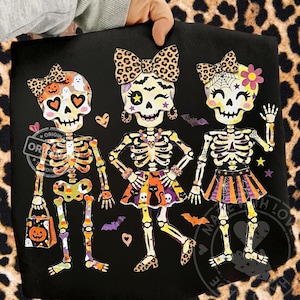 May include: A black t-shirt featuring a graphic of three skeletons with leopard print bows. The skeletons are decorated with Halloween motifs including pumpkins, bats, a black cat, and orange and purple striped skirts, set against a black background.
