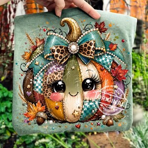 May include: A sage green t-shirt featuring a graphic of a cartoon pumpkin with large eyes, long eyelashes, and a smiling mouth. The pumpkin is decorated with a leopard print bow, pearls, autumn leaves, acorns, and stars, with the word ORIGINAL printed.