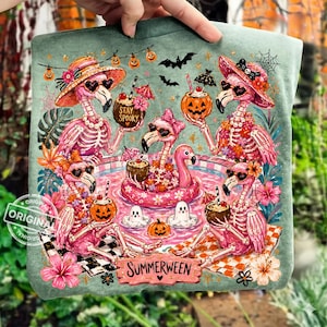 May include: A teal t-shirt featuring a whimsical illustration of skeleton flamingos in a pool, holding cocktails and jack-o'-lanterns. The design includes the text "Stay Spooky" and "Summerween", with bats and Halloween decorations.