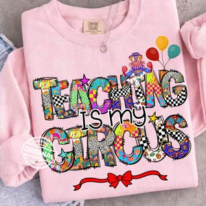 Może przedstawiać: Różowa bluza z napisem "TEACHING IS MY CIRCUS" w kolorowym, patchworkowym wzorze. Grafika klauna i balony znajdują się nad słowem "TEACHING". Czerwona wstążka z kokardą znajduje się na dole.