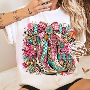 May include: A white t-shirt featuring a graphic print of two cowboy boots adorned with pink leopard print, teal checkered patterns, and a large bow. The design includes festive elements like poinsettias, wrapped gifts, and gold bells.