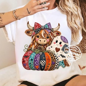 May include: A white t-shirt featuring a graphic of a brown Highland cow with a colorful patterned bow, sitting behind a large pumpkin decorated with various fabric textures and prints, accompanied by a small white ghost with a heart pattern.