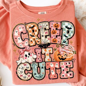May include: A coral-colored crewneck sweatshirt featuring a large graphic print with the text "CREEP IT CUTE". The design includes colorful patterned letters filled with ghosts, pumpkins, bats, spiderwebs, skulls, and candy corn motifs.