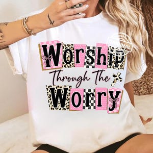 Könnte beinhalten: Weißes T-Shirt mit dem Schriftzug "Worship Through The Worry" in Schwarz, Rosa und Leopardenmuster. Das Design enthält eine Kreuzgrafik. Der Text ist in einer fetten, stilisierten Schriftart.