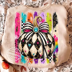 May include: A beige sweatshirt featuring a graphic of a pumpkin with a black and white harlequin pattern, adorned with a large striped bow, a turquoise gem, and gold glitter stem. The design includes colorful vertical brushstrokes and stars.
