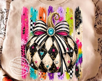 May include: A beige sweatshirt featuring a graphic of a pumpkin with a black and white harlequin pattern, adorned with a large striped bow, a turquoise gem, and gold glitter stem. The design includes colourful vertical brushstrokes and stars.