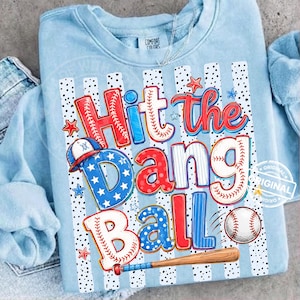 May include: Pale blue sweatshirt with a baseball-themed design. The graphic reads "Hit the Dang Ball" in red, white, and blue, with a baseball, bat, and stars. The sweatshirt has a crew neck and long sleeves.
