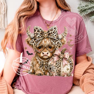 May include: A mauve short-sleeve t-shirt featuring a graphic of a Highland cow wearing a leopard print bow, heart-shaped glasses, and a pearl necklace. Next to the cow is a leopard print pumpkin with a pink bow, small stars, and a bat.