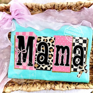 Puede incluir: Una camiseta azul claro con la palabra "Mama" en letras negras grandes. Cada letra está sobre un fondo con un patrón diferente, incluyendo cuadros rosas, estampado de leopardo y lunares. Un lazo rosa está en la "M" y la "a". Una cruz con estampado de leopardo está a la derecha.